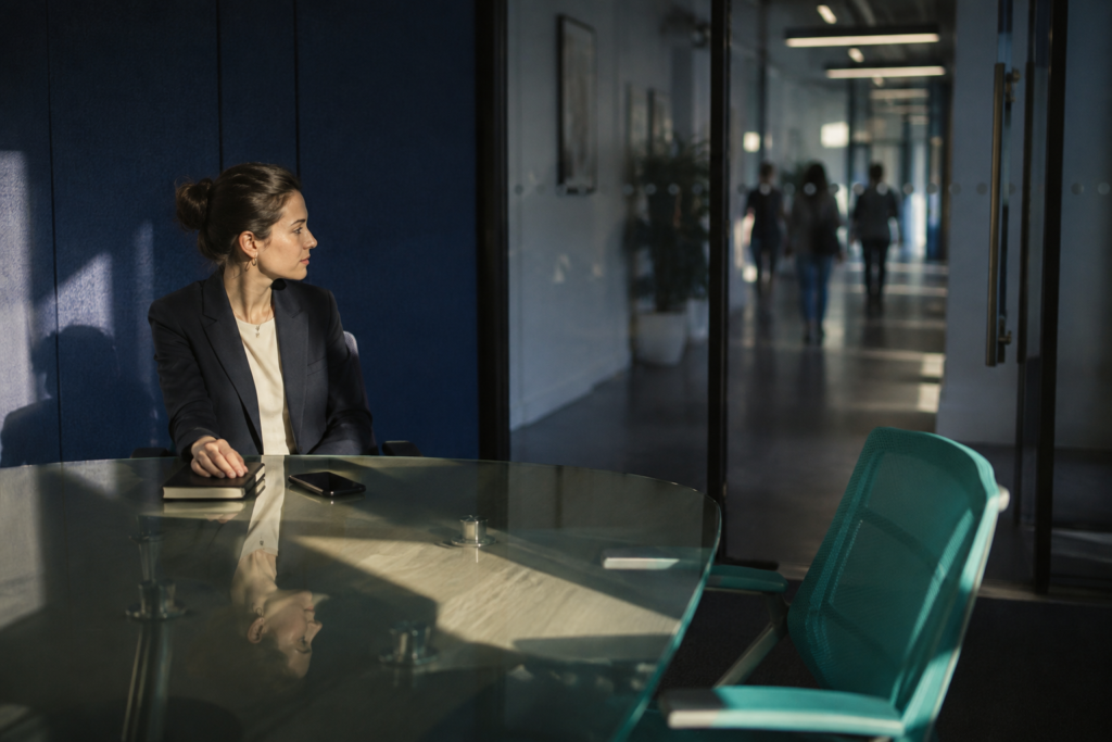 Employee waiting alone in a glass meeting room beside an empty chair, her manager's one-to-one not happening.